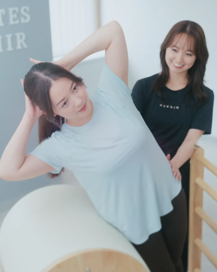 ラダーバレルに身体をあずけて体側を伸ばす女性と、そばで見守るトレーナー